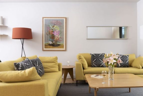 Bright living room with yellow sofas, a coffee table, a lamp, and floral artwork.