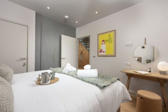 Modern bedroom with a double bed, light walls, and a wooden desk. A mirror and artwork decorate the space.