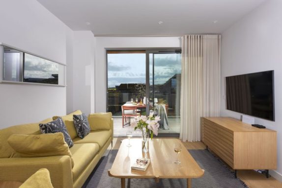 Modern living room with a yellow sofa, wooden coffee table, TV, and balcony access.