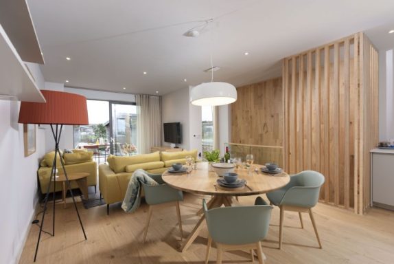 Modern dining area with a wooden table, green chairs, and a cosy living room in the background.